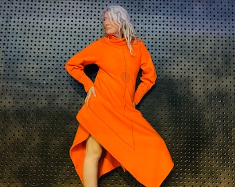 Op de afbeelding: Een vrouw draagt een lange, asymmetrische oranje jurk met een geometrisch ontwerp. De jurk heeft een hoge halslijn en lange mouwen. De vrouw staat voor een zwarte metalen muur met een patroon van gaten.