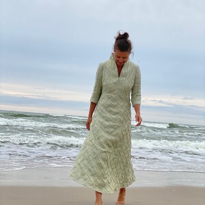May include: A woman wearing a light green maxi dress with a textured pattern walks on a sandy beach. The dress has a V-neckline and three-quarter sleeves.