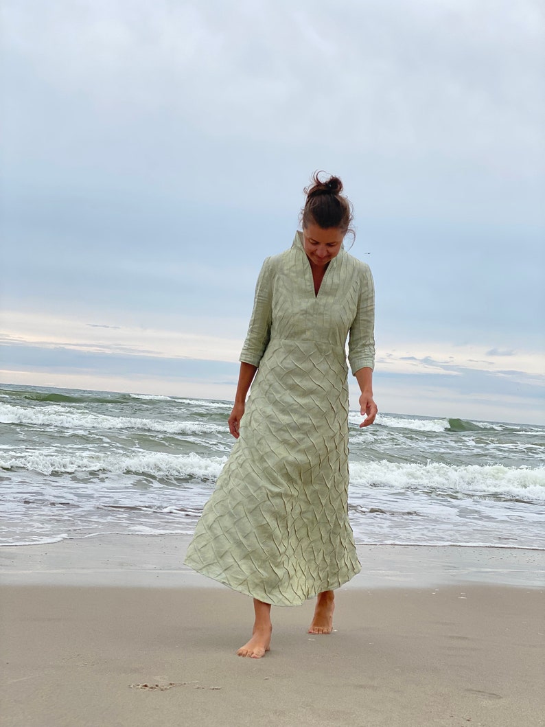 May include: A woman wearing a light green maxi dress with a textured pattern walks on a sandy beach. The dress has a V-neckline and three-quarter sleeves.