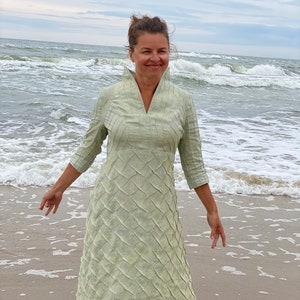 May include: A woman wearing a light green, long-sleeved dress with a textured pattern stands on a sandy beach with waves crashing in the background.