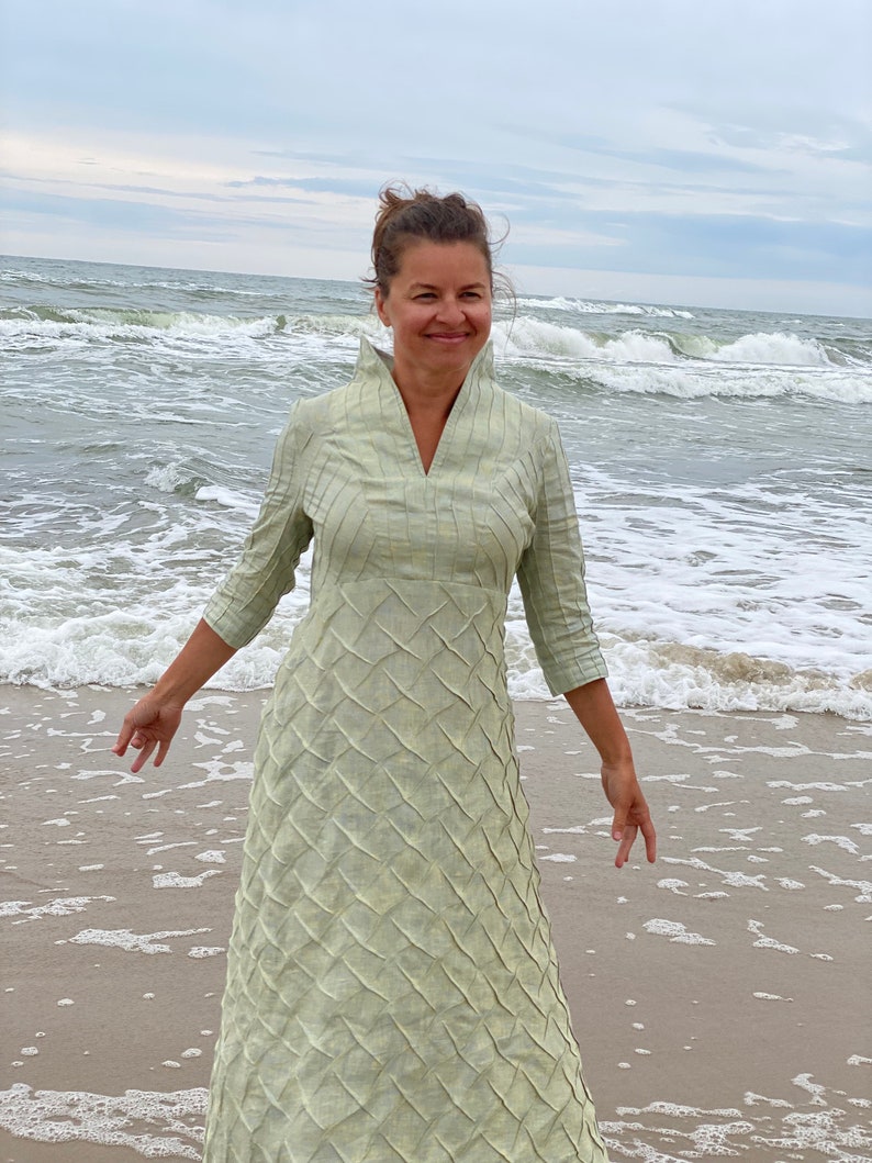 May include: A woman wearing a light green, long-sleeved dress with a textured pattern stands on a sandy beach with waves crashing in the background.