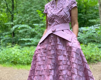 May include: A woman wearing a long, pink dress with a geometric pattern. The dress has a high collar and short sleeves. The woman is standing in a forest setting.