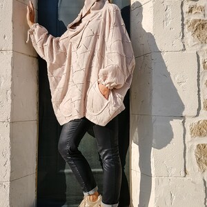 May include: A woman wearing a light pink, textured, oversized shirt with a high collar and black skinny jeans. She is wearing gold sneakers and sunglasses.