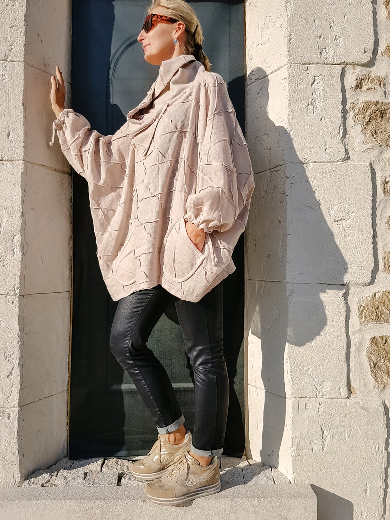 May include: A woman wearing a light pink, textured, oversized shirt with a high collar and black skinny jeans. She is wearing gold sneakers and sunglasses.