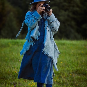 Puede incluir: Una persona con sombrero y abrigo azules, sosteniendo una cámara negra en un campo de hierba verde y flores amarillas. El abrigo tiene un diseño recortado único. La persona lleva un vestido azul y pantalones azules.