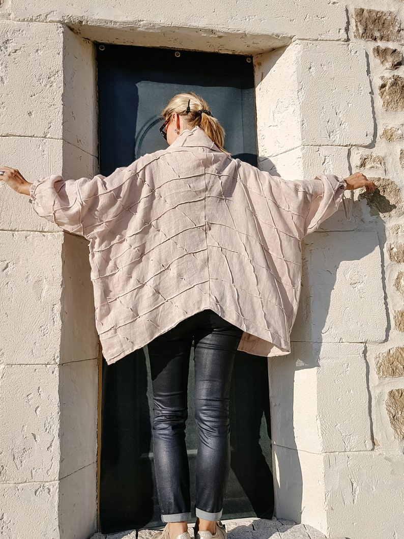 May include: A woman wearing a light pink, oversized, textured linen shirt with black skinny jeans. She is standing in front of a doorway with her arms outstretched.