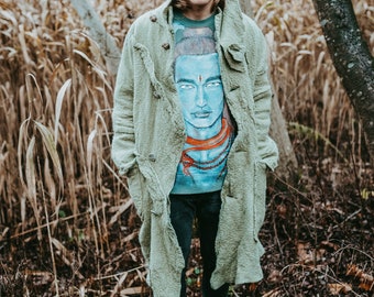 May include: A person wearing a light green coat over a blue graphic t-shirt featuring a blue face with a red dot and necklace. The person is also wearing black pants and white sneakers. The background is a forest.