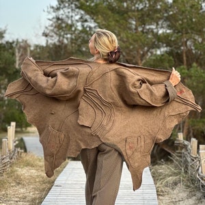 Puede incluir: Un abrigo de lana marrón con un diseño asimétrico, con cortes y bolsillos únicos. El abrigo se lleva abierto, revelando pantalones marrones estampados. El fondo incluye una pasarela de madera y árboles.