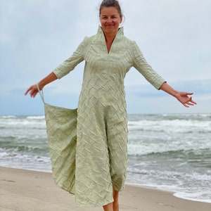 May include: A woman wearing a light green, long-sleeved dress with a textured pattern, standing on a sandy beach with the ocean in the background.