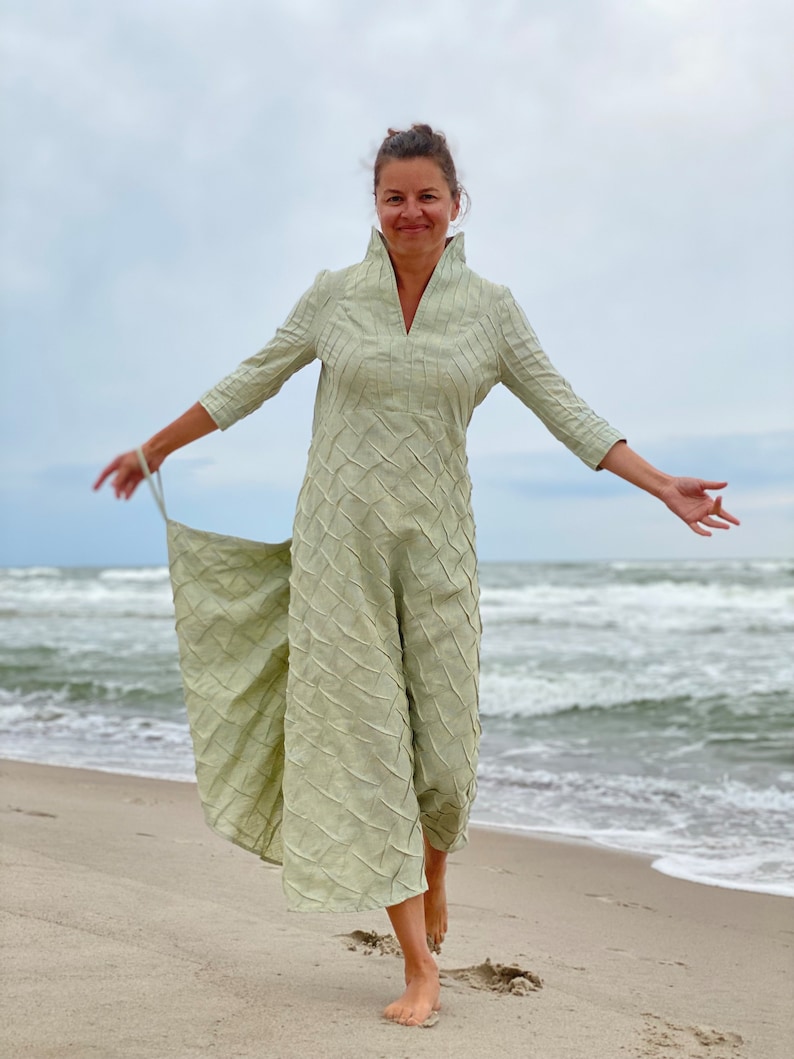 May include: A woman wearing a light green, long-sleeved dress with a textured pattern, standing on a sandy beach with the ocean in the background.