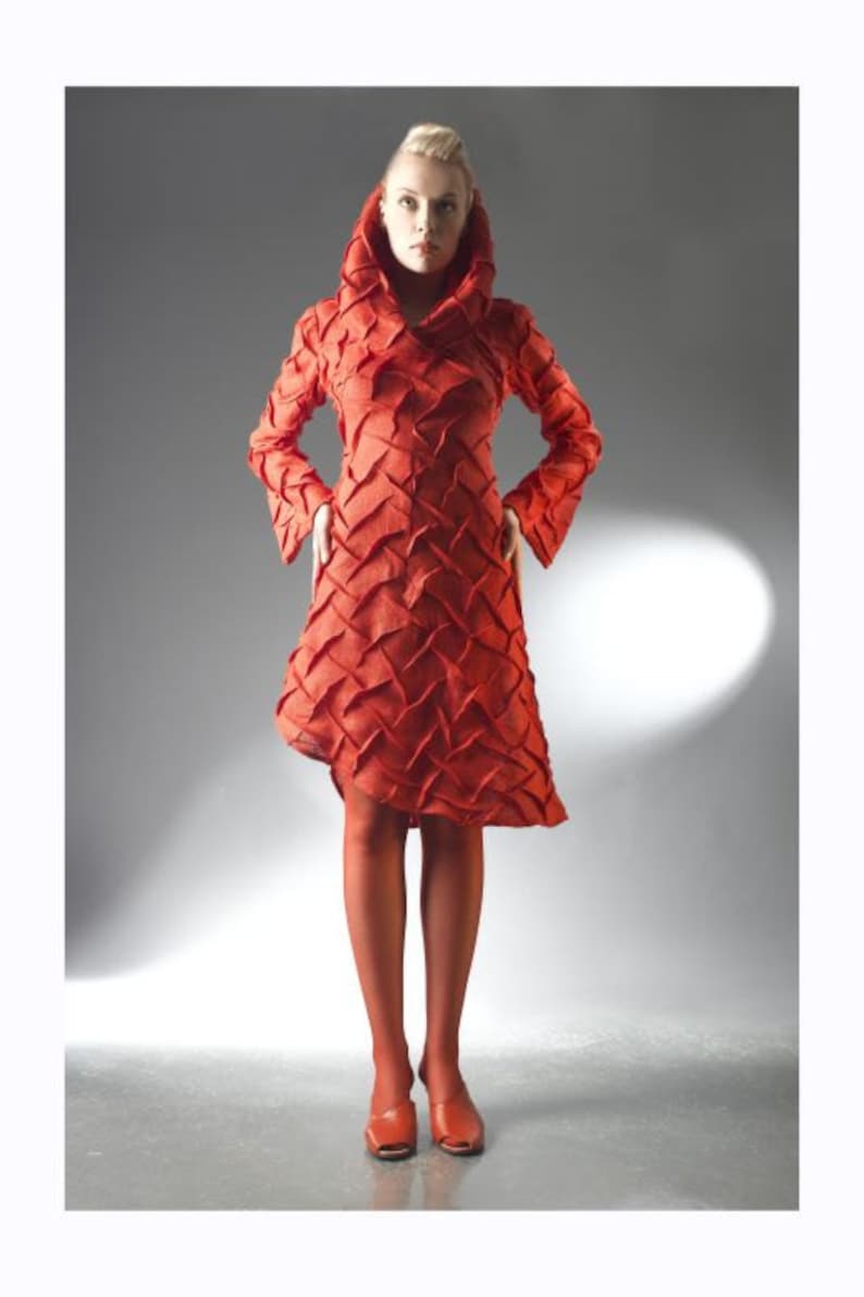 Peut inclure: Une femme portant une robe rouge &agrave; capuche textur&eacute;e avec un motif g&eacute;om&eacute;trique. Elle est debout sur un fond gris et porte des chaussures rouges.