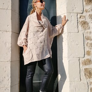 May include: A woman wearing a light brown, loose-fitting, textured top with a cowl neck, black skinny jeans, and gold sneakers. She is standing in a doorway with a white stone wall on one side and a dark blue door on the other.