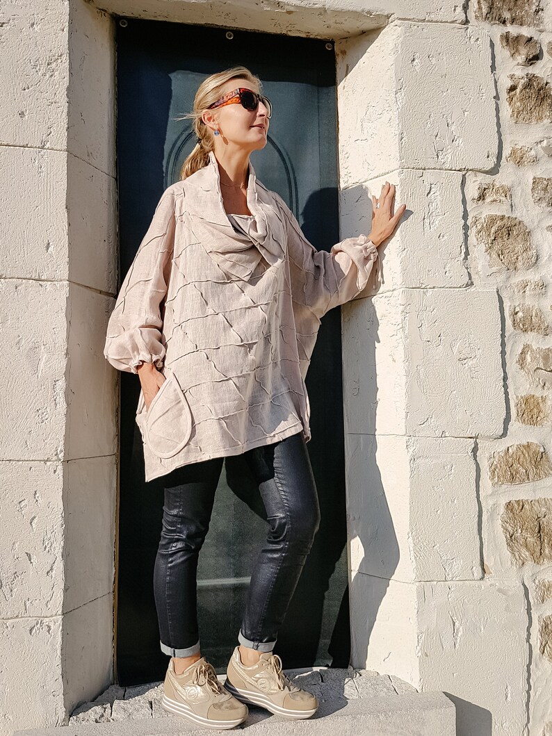 May include: A woman wearing a light brown, loose-fitting, textured top with a cowl neck, black skinny jeans, and gold sneakers. She is standing in a doorway with a white stone wall on one side and a dark blue door on the other.