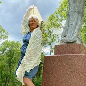 Puede incluir: Una mujer con un chal blanco y un vestido de mezclilla azul se para frente a una estatua de &aacute;ngel de piedra. La estatua est&aacute; sobre un pedestal de piedra con escalones. La mujer sostiene el chal por encima de su cabeza. El fondo es un &aacute;rea de hierba verde con &aacute;rboles.