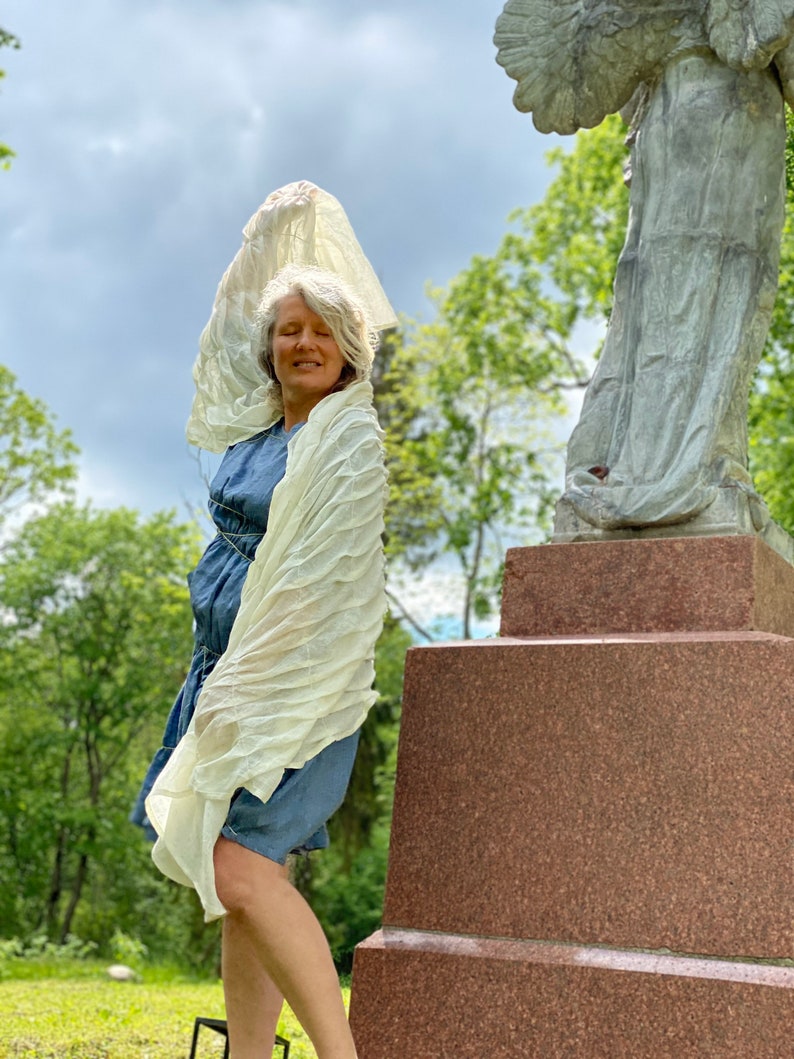 Puede incluir: Una mujer con un chal blanco y un vestido de mezclilla azul se para frente a una estatua de &aacute;ngel de piedra. La estatua est&aacute; sobre un pedestal de piedra con escalones. La mujer sostiene el chal por encima de su cabeza. El fondo es un &aacute;rea de hierba verde con &aacute;rboles.