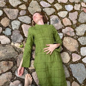 以下が含まれることがあります： 緑色のキルティングドレスを着た女性が石畳の道に横たわっています。ドレスはハイネックで長袖です。