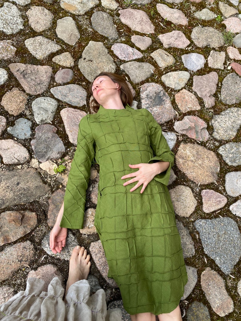 以下が含まれることがあります： 緑色のキルティングドレスを着た女性が石畳の道に横たわっています。ドレスはハイネックで長袖です。
