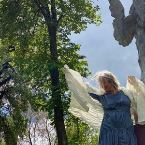 Puede incluir: Una mujer con un vestido azul con tela blanca drapeada sobre sus brazos, de pie en un bosque con una gran estatua de &aacute;ngel de piedra en el fondo.