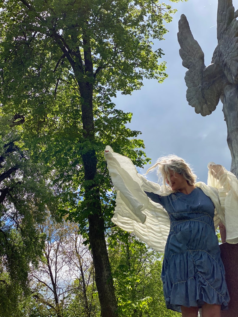 Puede incluir: Una mujer con un vestido azul con tela blanca drapeada sobre sus brazos, de pie en un bosque con una gran estatua de &aacute;ngel de piedra en el fondo.