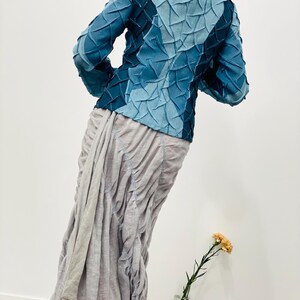 May include: A blue denim jacket with a unique textured design. The jacket has a high collar and long sleeves. The model is wearing a grey skirt.