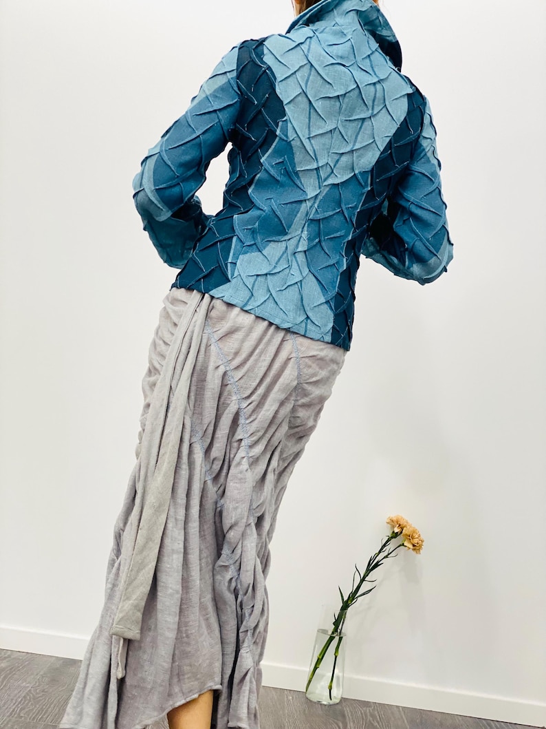 May include: A blue denim jacket with a unique textured design. The jacket has a high collar and long sleeves. The model is wearing a grey skirt.