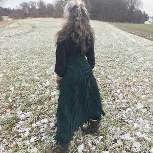 May include: A woman wearing a dark green, long, asymmetrical skirt with a textured design. The skirt is made of a soft, flowing fabric. She is standing in a snowy field with trees in the background.