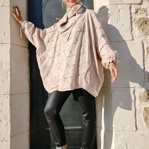 May include: A woman wearing a light pink, textured, oversized sweater with a cowl neck, black leather pants, and beige sneakers. She is standing next to a door.