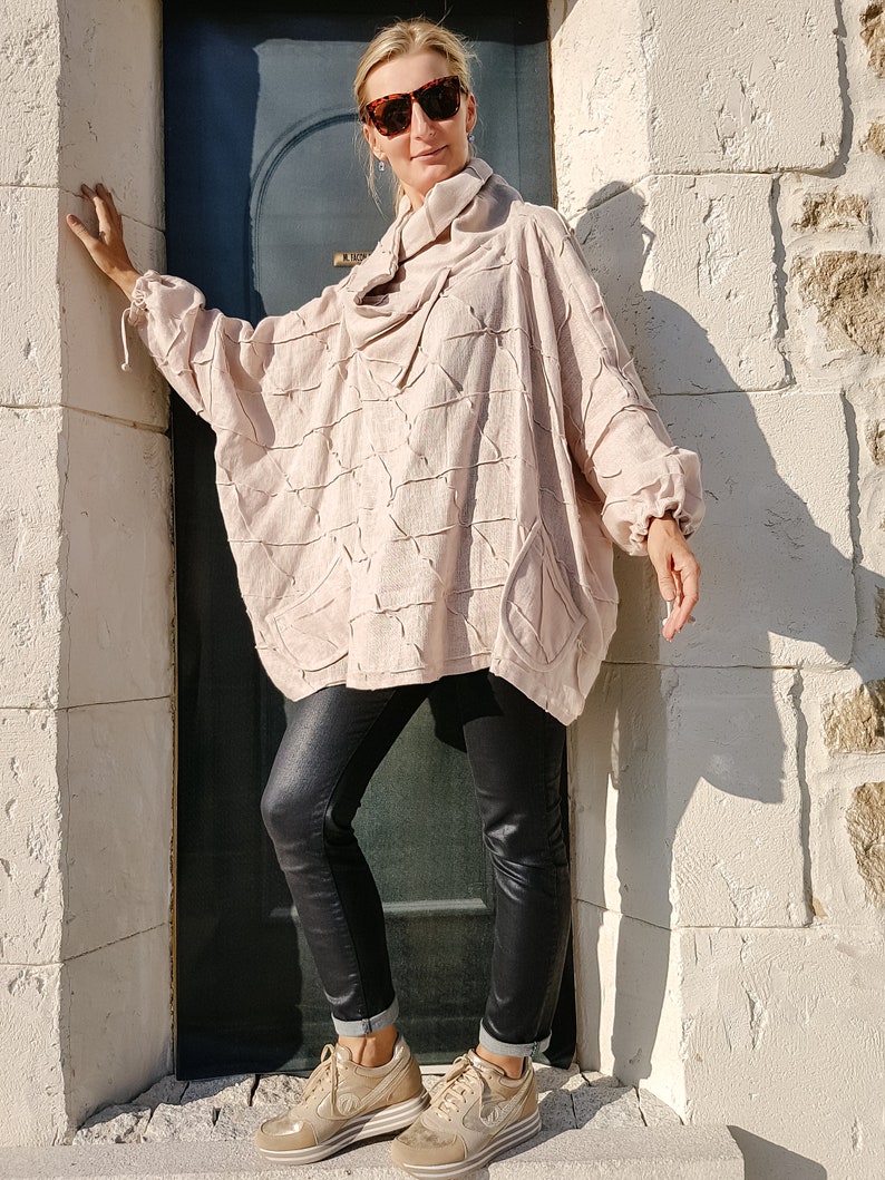 May include: A woman wearing a light pink, textured, oversized sweater with a cowl neck, black leather pants, and beige sneakers. She is standing next to a door.