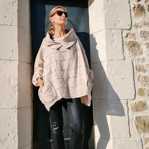 May include: A woman wearing a light pink, textured, oversized shirt with a cowl neck and black jeans. She is wearing sunglasses and standing in front of a white brick wall.