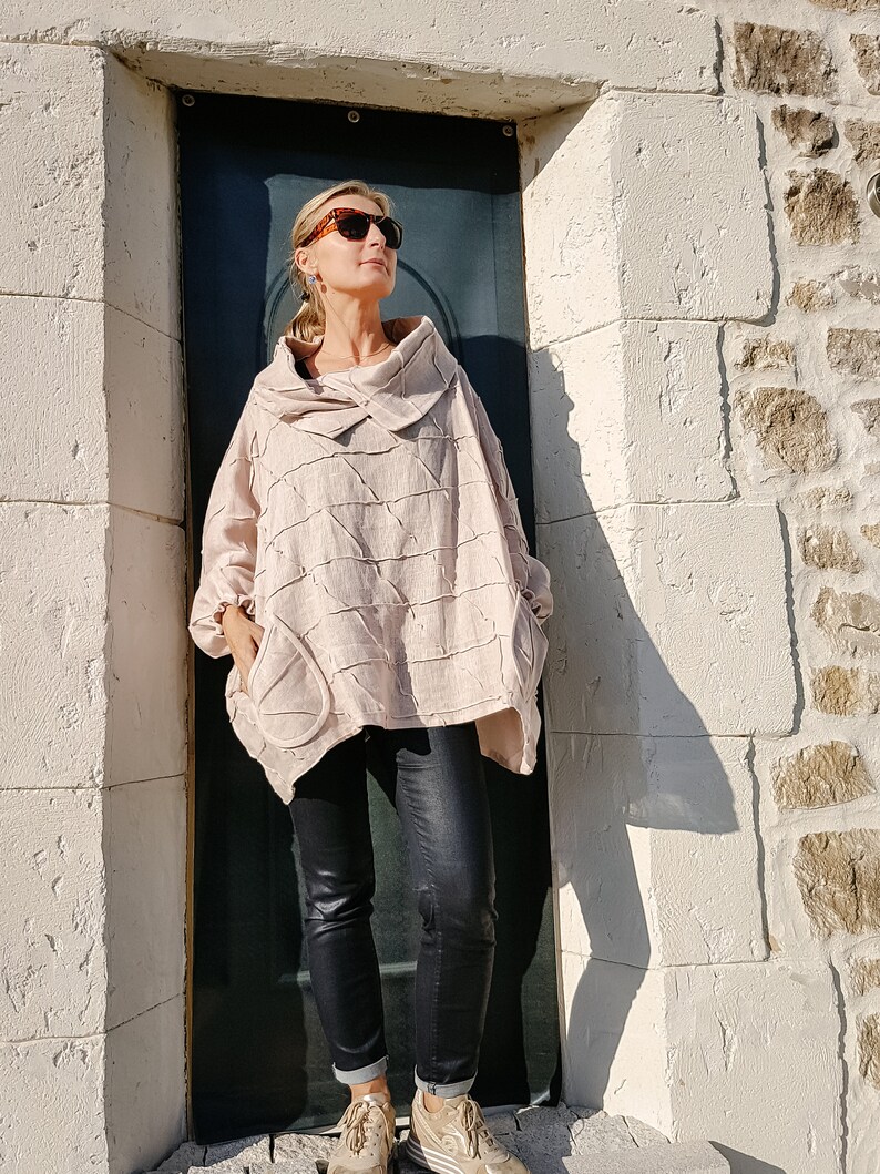 May include: A woman wearing a light pink, textured, oversized shirt with a cowl neck and black jeans. She is wearing sunglasses and standing in front of a white brick wall.