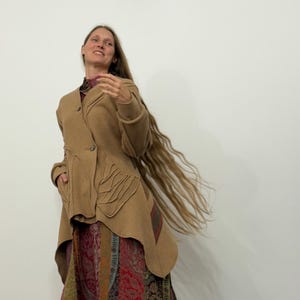 Puede incluir: Chaqueta asimétrica de color beige con detalles de costura únicos y cierre de botón. La chaqueta se combina con una falda larga estampada en tonos rojos, verdes y dorados. La persona tiene cabello largo y suelto.