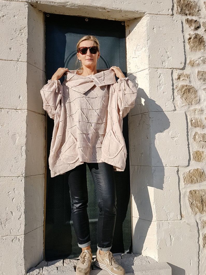 May include: A woman wearing a light pink, oversized,  crinkled linen top with a cowl neck and long sleeves. She is wearing black skinny jeans and tan sneakers.