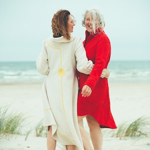 May include: Two people wearing long coats on a beach. One coat is cream-colored with yellow accents, the other is red. The coats have unique designs and are knee-length. The background shows the ocean and sand.