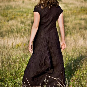 May include: A woman wearing a black, textured dress walks through a field of long grass.