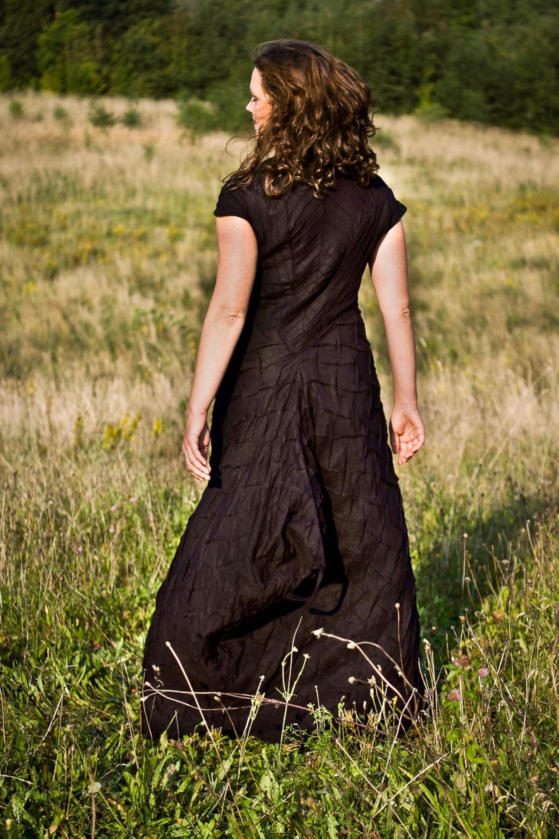 May include: A woman wearing a black, textured dress walks through a field of long grass.