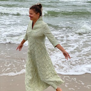May include: A woman wearing a light green linen dress with a textured pattern walks on a sandy beach with waves crashing in the background.