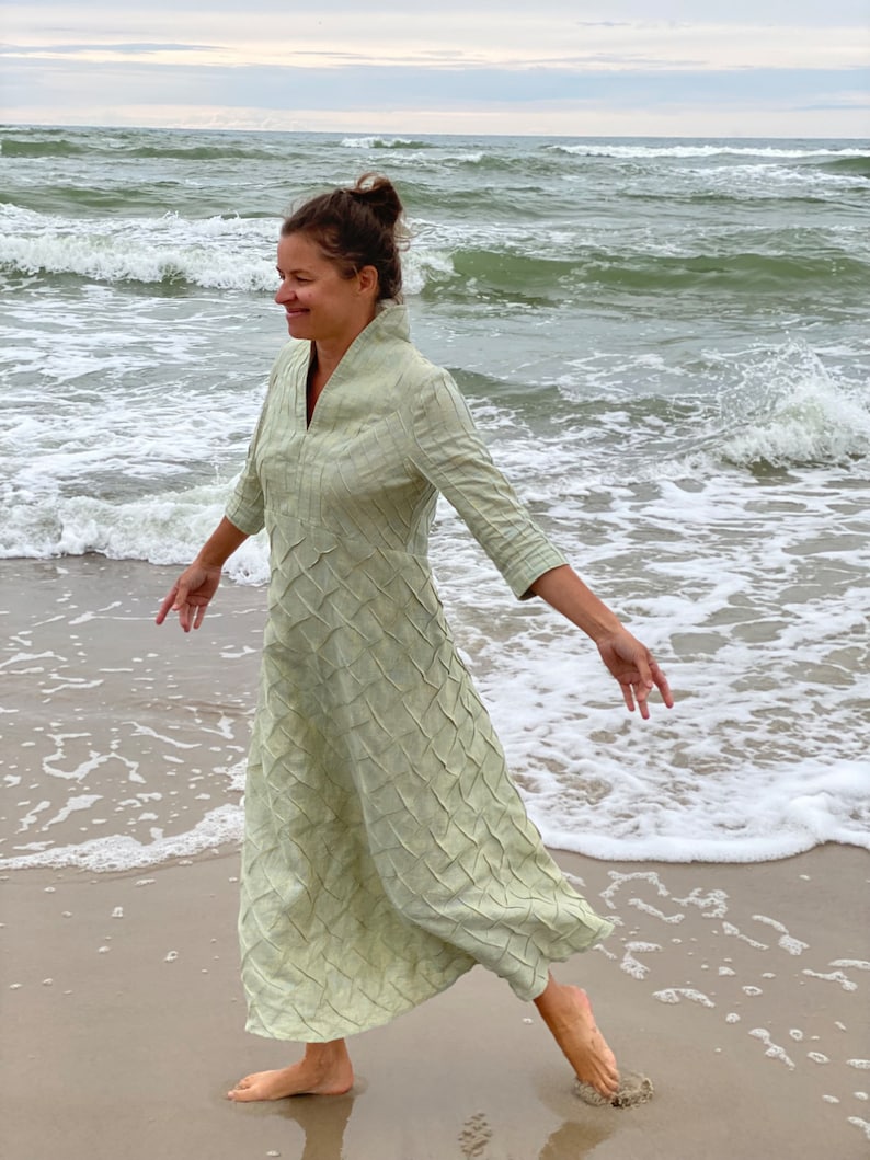 May include: A woman wearing a light green linen dress with a textured pattern walks on a sandy beach with waves crashing in the background.