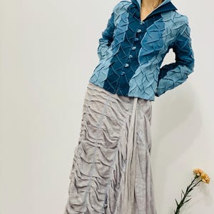 May include: A blue denim jacket with a textured, quilted design. The jacket has a collar, long sleeves, and a button closure. The model is wearing a light grey skirt.