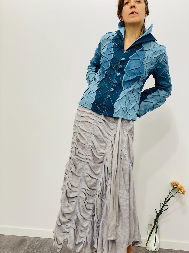 May include: A blue denim jacket with a textured, quilted design. The jacket has a collar, long sleeves, and a button closure. The model is wearing a light grey skirt.