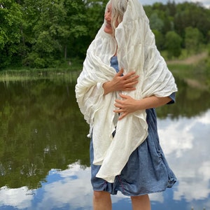 Puede incluir: Una mujer con un chal de lino blanco sobre un vestido de mezclilla azul, de pie junto a un lago con &aacute;rboles verdes en el fondo.