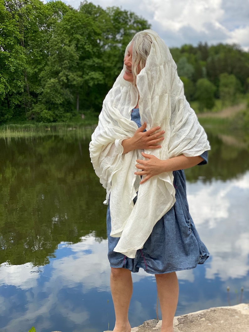 Puede incluir: Una mujer con un chal de lino blanco sobre un vestido de mezclilla azul, de pie junto a un lago con &aacute;rboles verdes en el fondo.