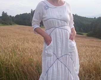 May include: A white linen dress with blue stitching and a loose, flowing silhouette. The dress has a round neckline and three-quarter sleeves. The model is standing in a field of tall grass.