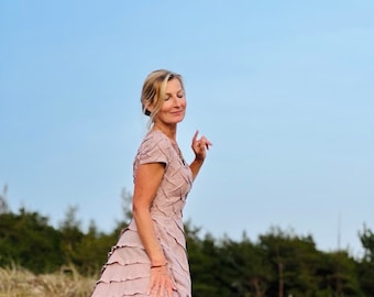 May include: A woman in a long, ruffled, blush-colored dress stands on a path with a wooden fence and dry grass in the background.