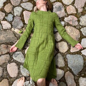以下が含まれることがあります： 緑色の格子柄のドレスを着た女性が石畳の道に横たわっています。彼女は目を閉じ、腕を伸ばしています。