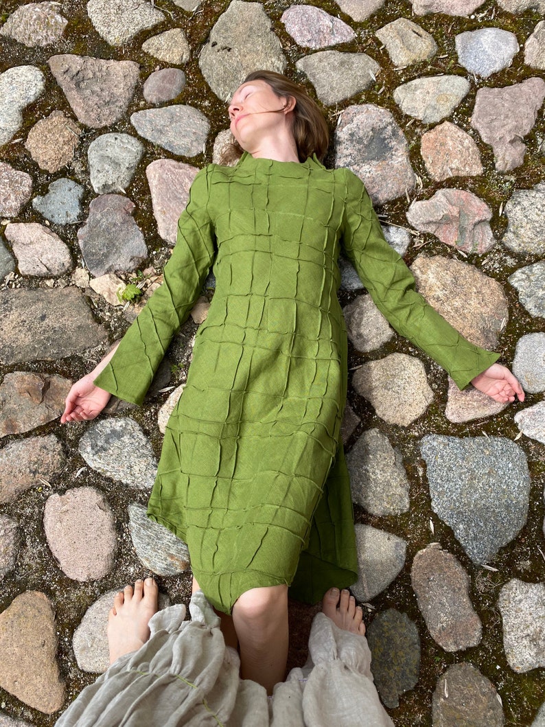 以下が含まれることがあります： 緑色の格子柄のドレスを着た女性が石畳の道に横たわっています。彼女は目を閉じ、腕を伸ばしています。