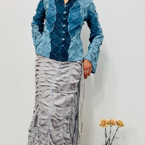May include: A blue and teal textured jacket with a button-up front and long sleeves. The jacket is paired with a long, flowing, grey skirt.