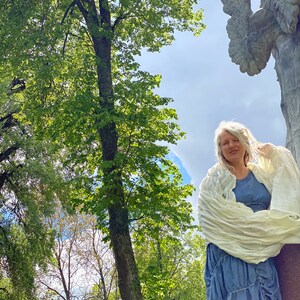 Puede incluir: Una mujer con cabello gris se para frente a una estatua de &aacute;ngel de piedra con alas. Lleva una chalina blanca sobre un vestido azul. La estatua es parcialmente visible en el fondo. La mujer est&aacute; de pie en un parque con &aacute;rboles y un cielo azul.