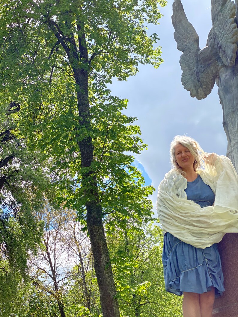 Puede incluir: Una mujer con cabello gris se para frente a una estatua de &aacute;ngel de piedra con alas. Lleva una chalina blanca sobre un vestido azul. La estatua es parcialmente visible en el fondo. La mujer est&aacute; de pie en un parque con &aacute;rboles y un cielo azul.