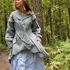 Puede incluir: Una chaqueta gris texturizada, asimétrica, con cuello alto y cierre de botones. La chaqueta se lleva sobre una falda azul claro con diseño plisado.