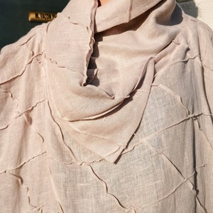 May include: A light beige linen poncho with a high, gathered neckline. The fabric has a textured, woven look and is stitched with a decorative, uneven seam.